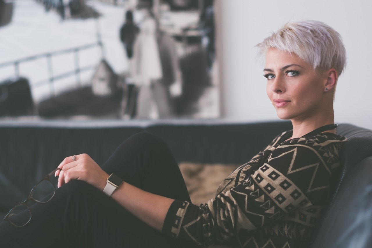 A mom sitting on a couch, wearing diamond stud earrings, a rind, and a smartwatch while holding her glasses in one hand