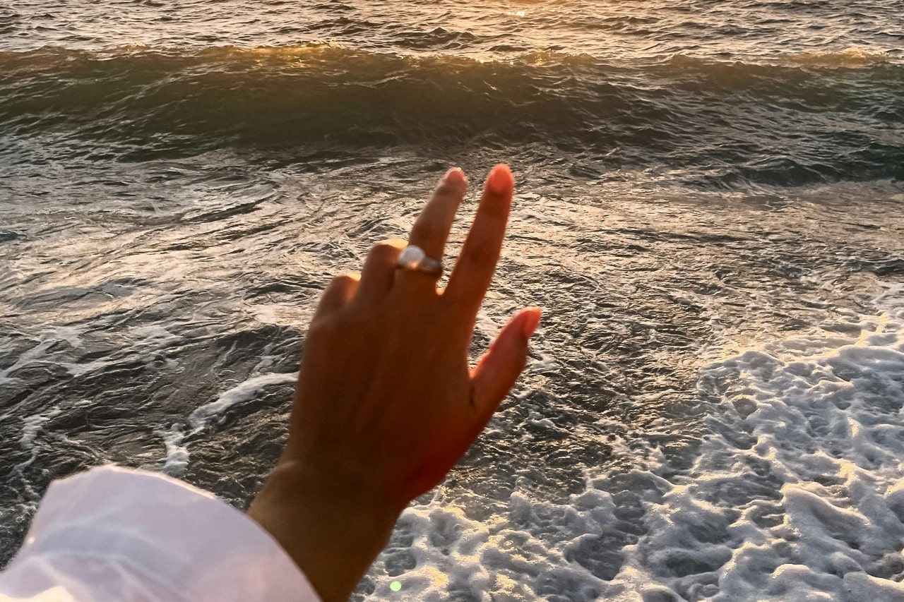 A woman extends her hand toward the ocean, wearing a large diamond ring.