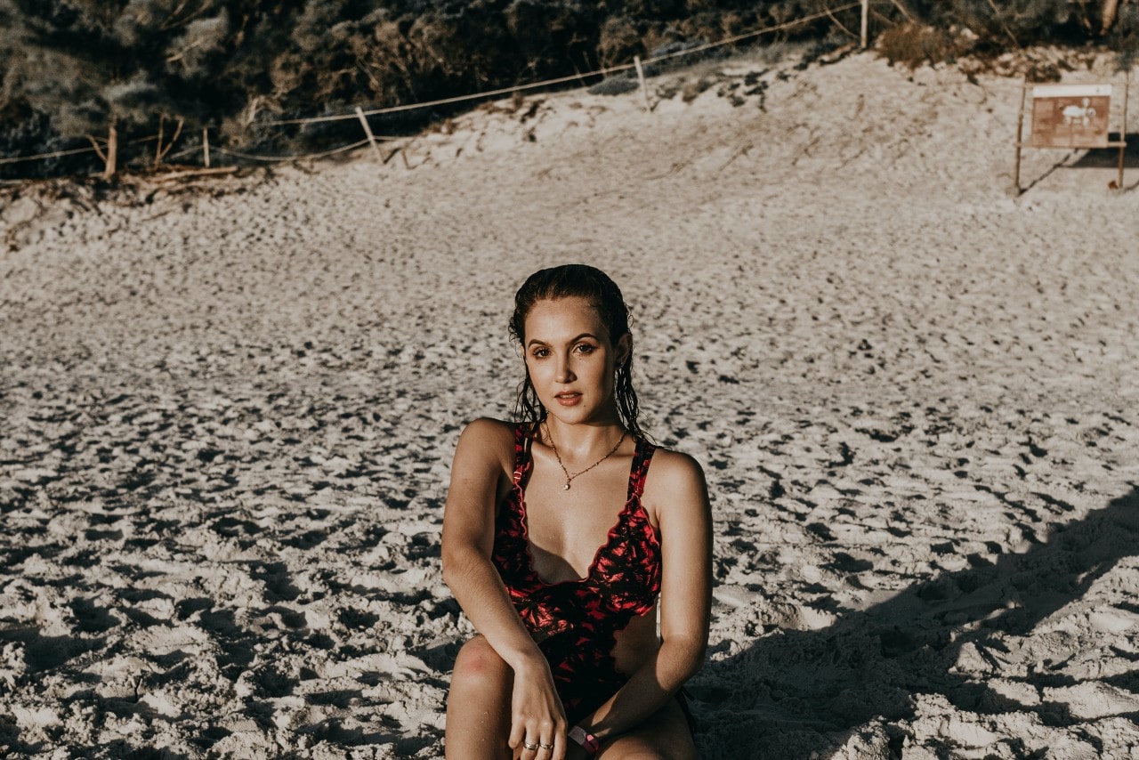 a woman sitting in the sand wearing a swimsuit and a pendant necklace